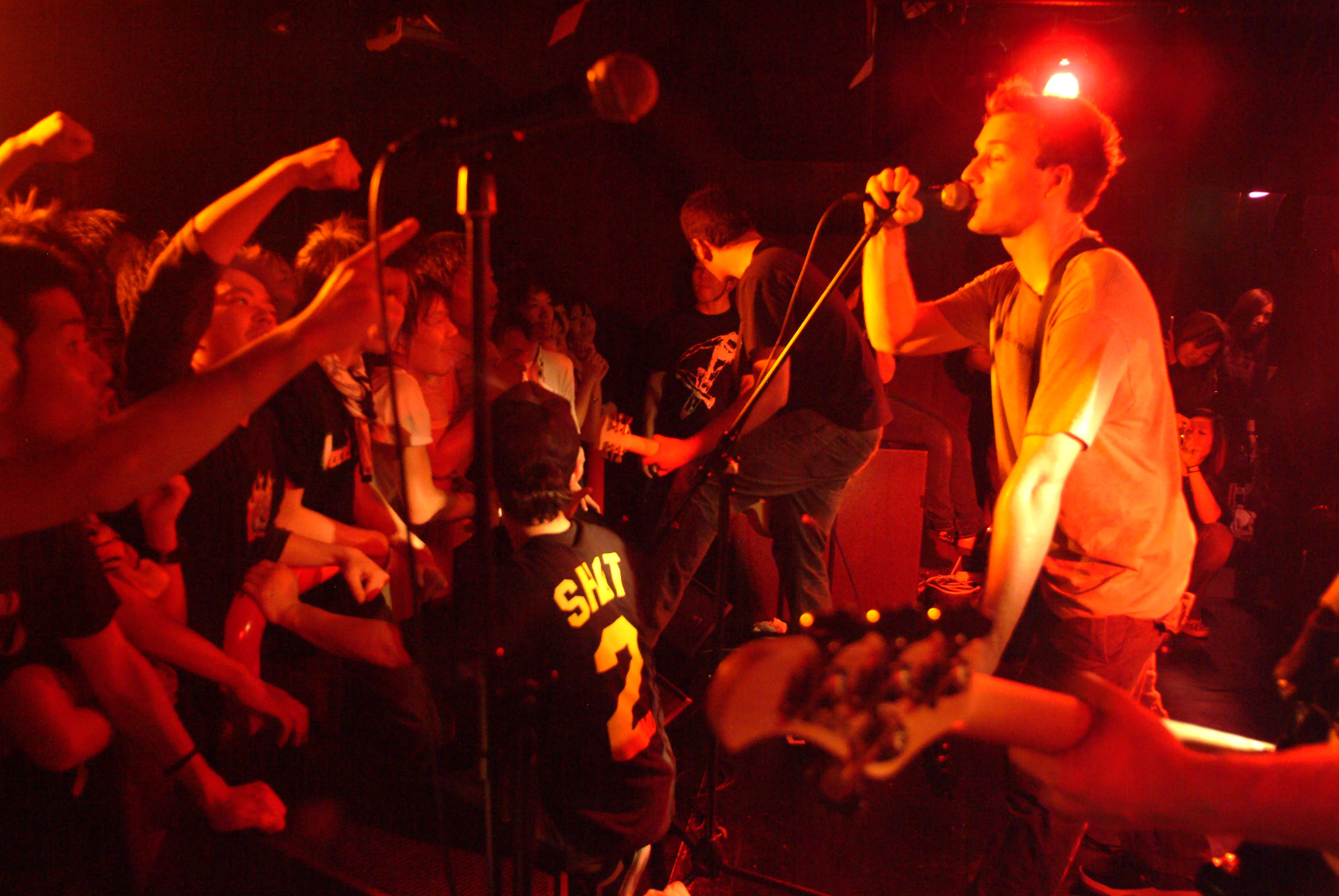 SecondShot singer at the mic in a red-lit club while fans in the front row throw fists in the air