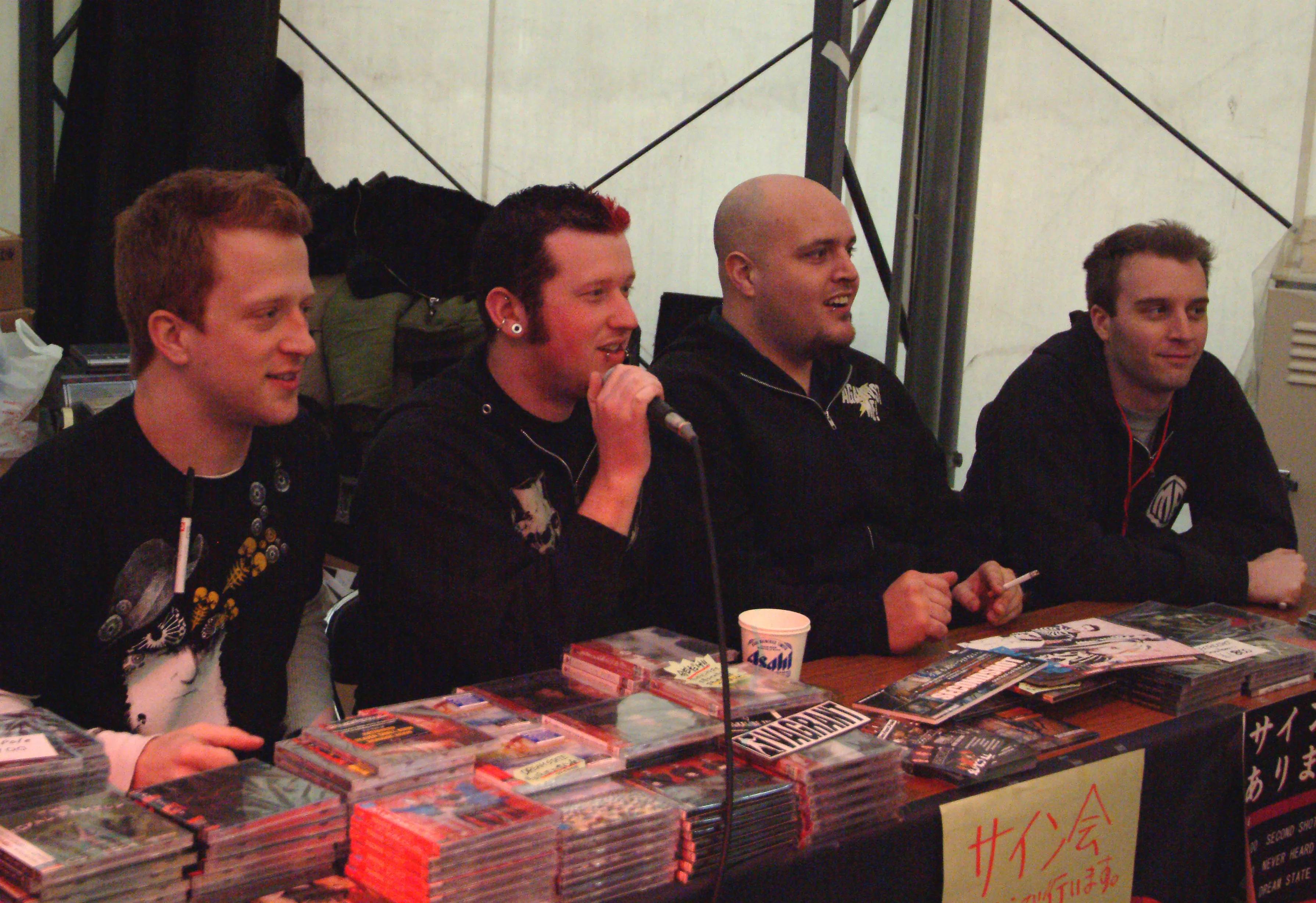 Four SecondShot members at a merch table stacked with CDs during a signing session in Japan