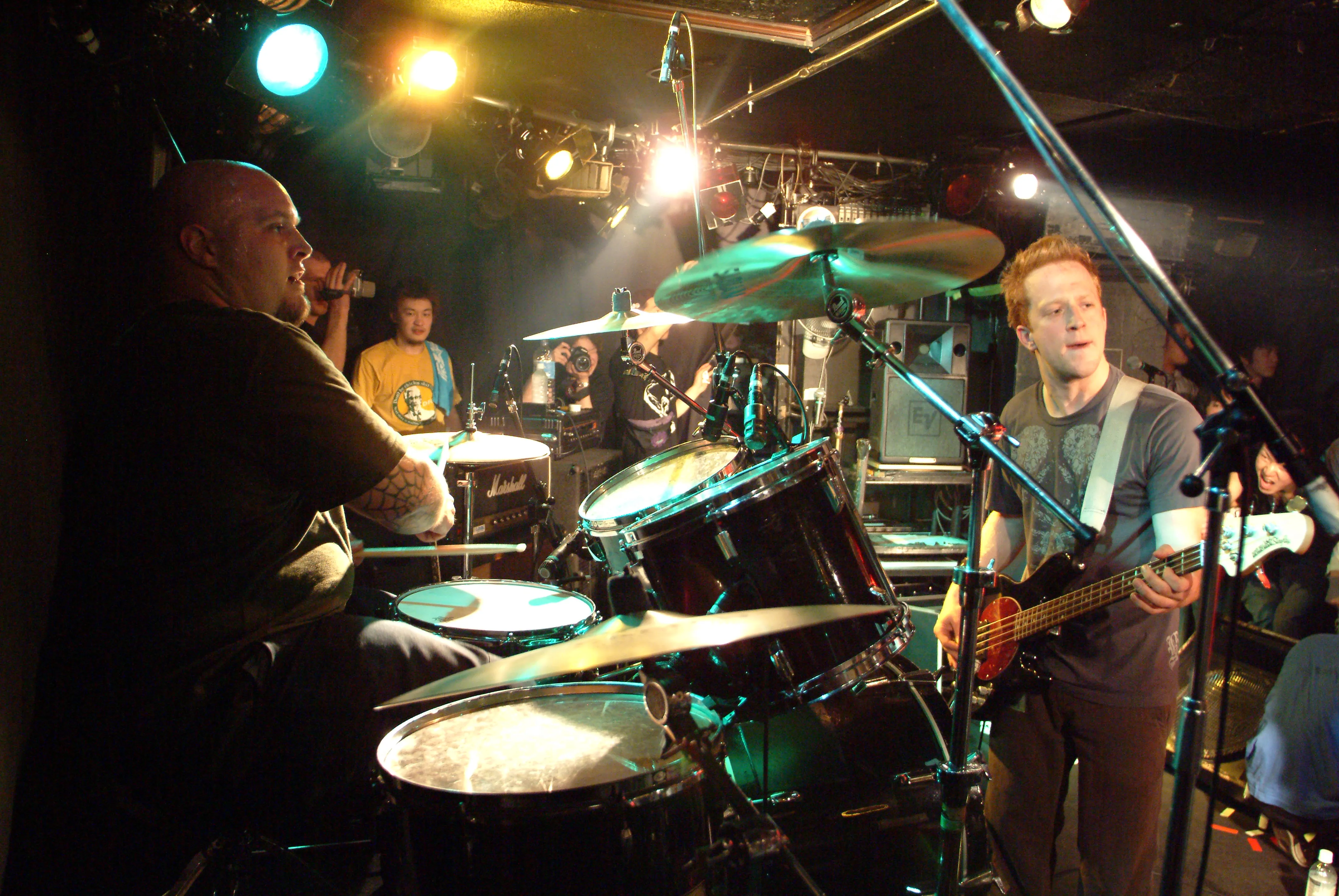 SecondShot drummer and bassist playing on a cramped club stage while fans film from behind the kit