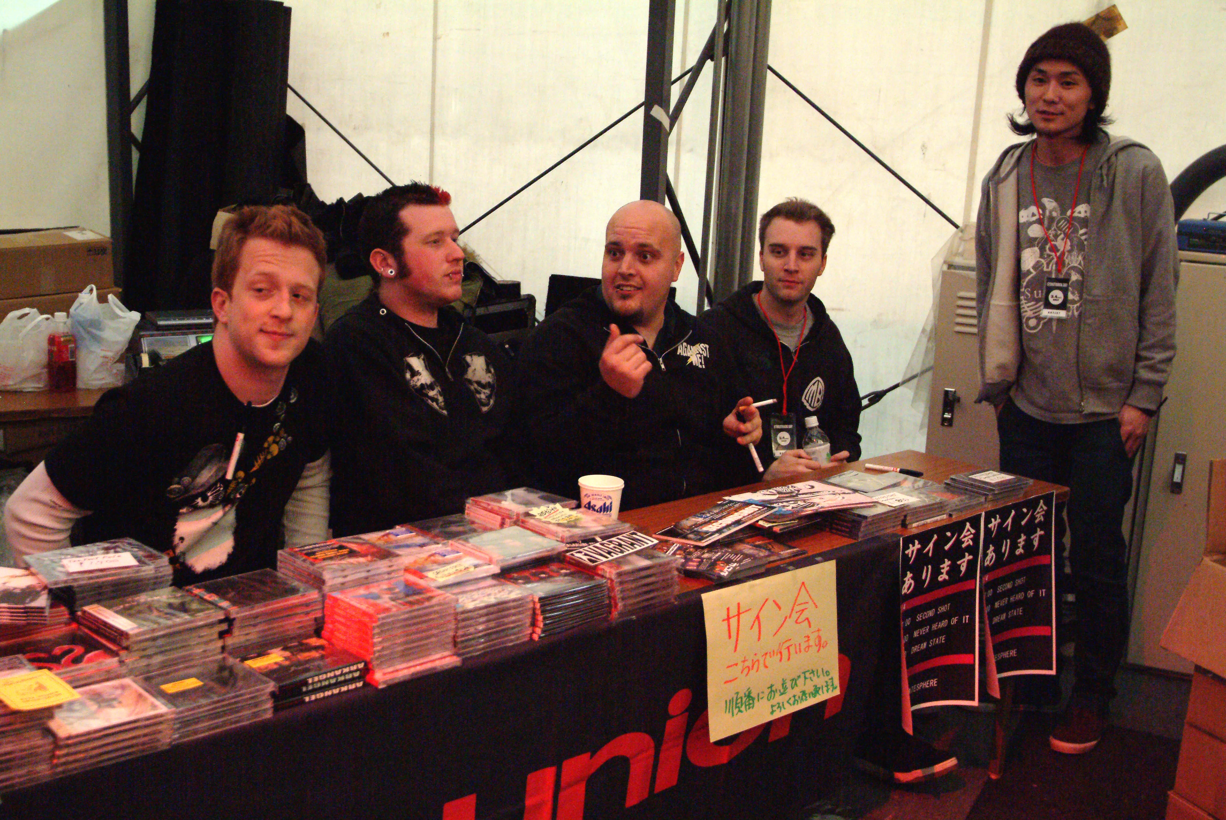 SecondShot at the merch table with a friend standing beside them, signing session signs on the table